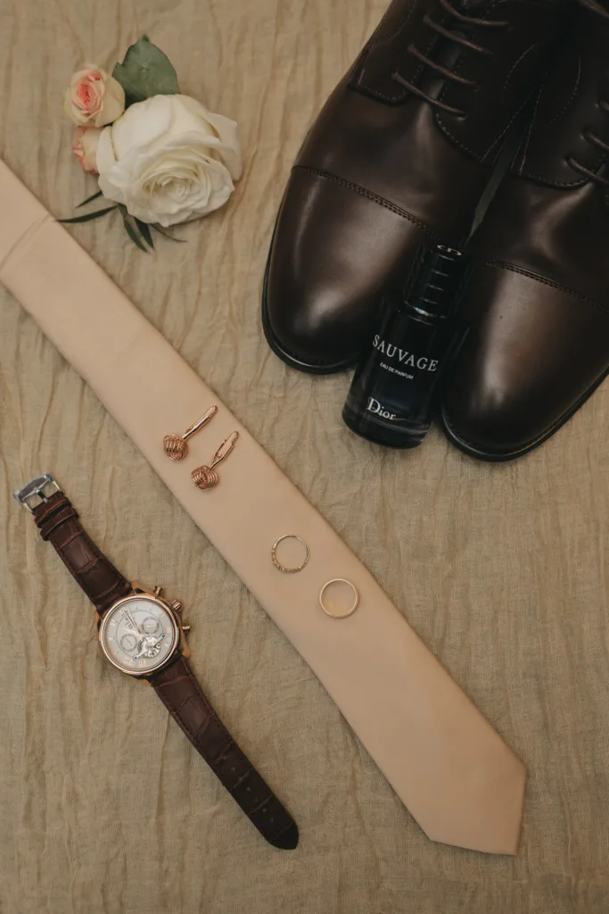 A flat lay of wedding accessories on beige fabric: a cream-colored tie, brown leather dress shoes, Dior Sauvage cologne, brown leather wristwatch, two rose gold cufflinks, two rings, and a white rose—perfect inspiration for candid wedding photography. © Aimee Lince Photography - Wedding photographer in Lincolnshire, Yorkshire & Nottinghamshire