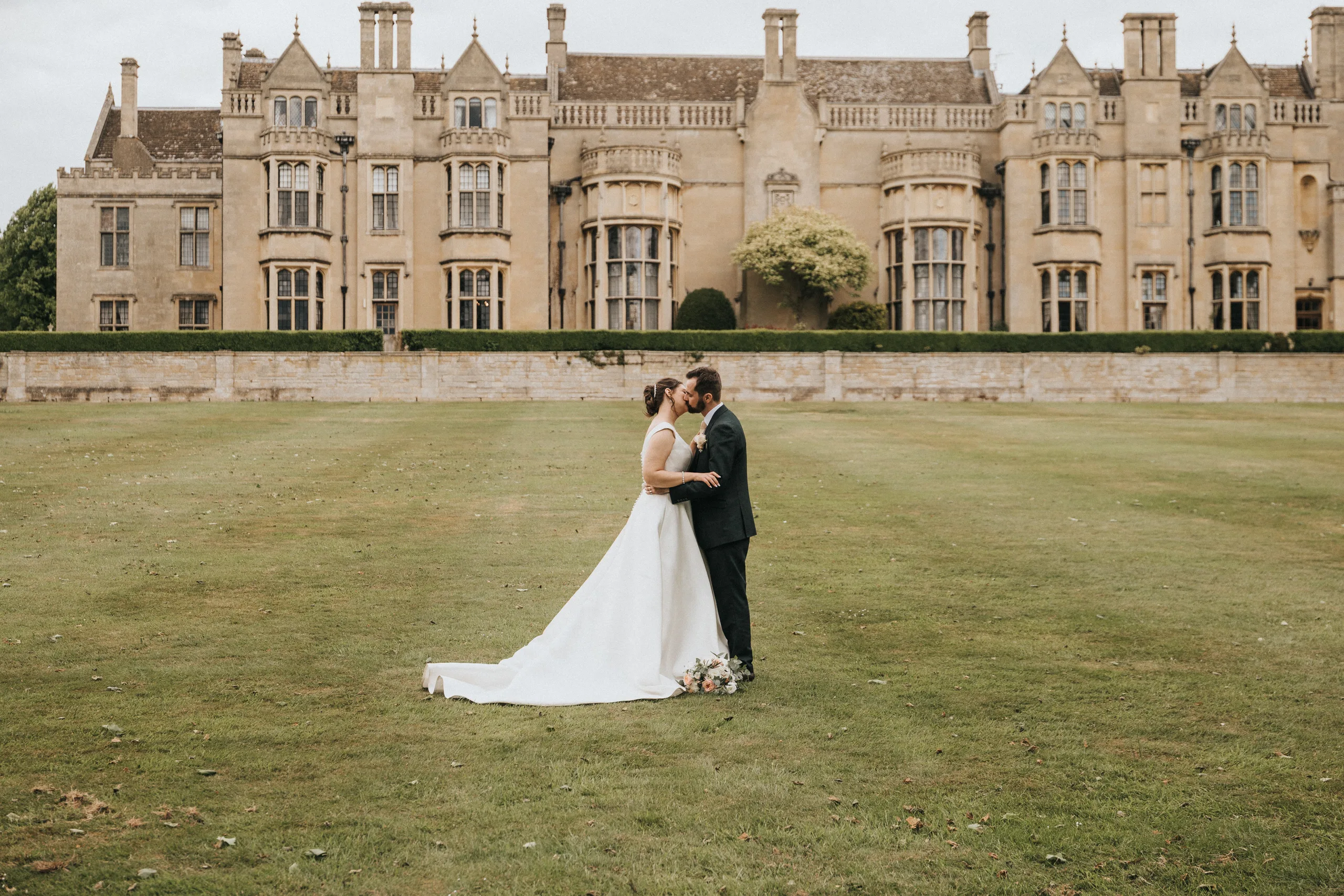 katie-adair-wedding-photography-rushton-hall-kettering-northamptonshire-706-of-1141-©-aimee-lince-photography-05-06-2025 A bride and groom stand facing each other on a large green lawn at Rushton Hall, gently touching foreheads. The bride wears a white gown with a long train, while the groom wears a dark suit by the grand historic manor—captured by an East Midlands wedding photographer. © Aimee Lince Photography - Wedding photographer in Lincolnshire, Yorkshire & Nottinghamshire