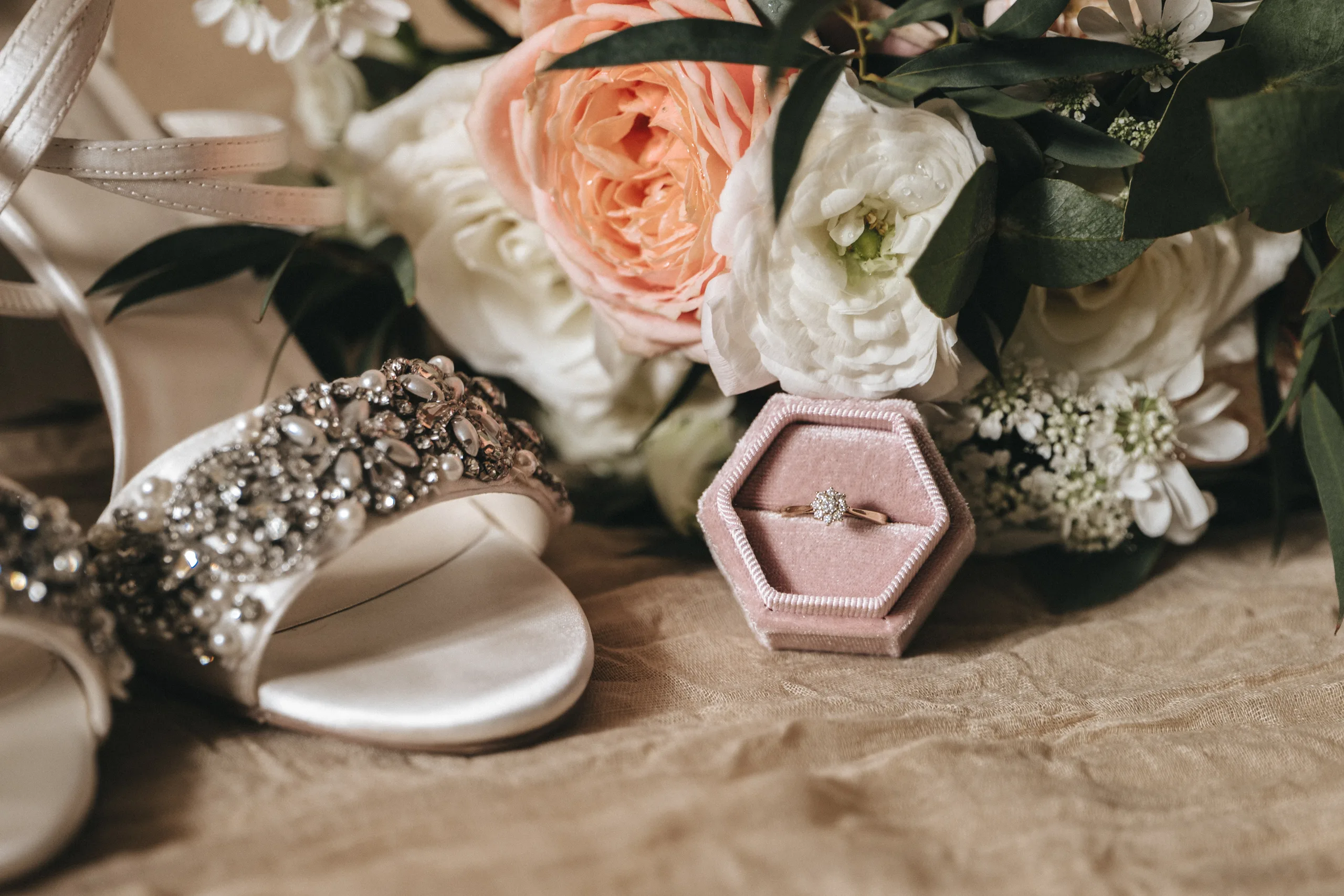katie-adair-wedding-photography-rushton-hall-kettering-northamptonshire-71-of-1141-©-aimee-lince-photography-02-06-2025 A close-up of a jeweled white bridal shoe, a diamond engagement ring in a pink hexagonal box, and a bouquet of white and peach flowers with greenery, all arranged on soft beige fabric—perfect inspiration for candid wedding photography. © Aimee Lince Photography - Wedding photographer in Lincolnshire, Yorkshire & Nottinghamshire