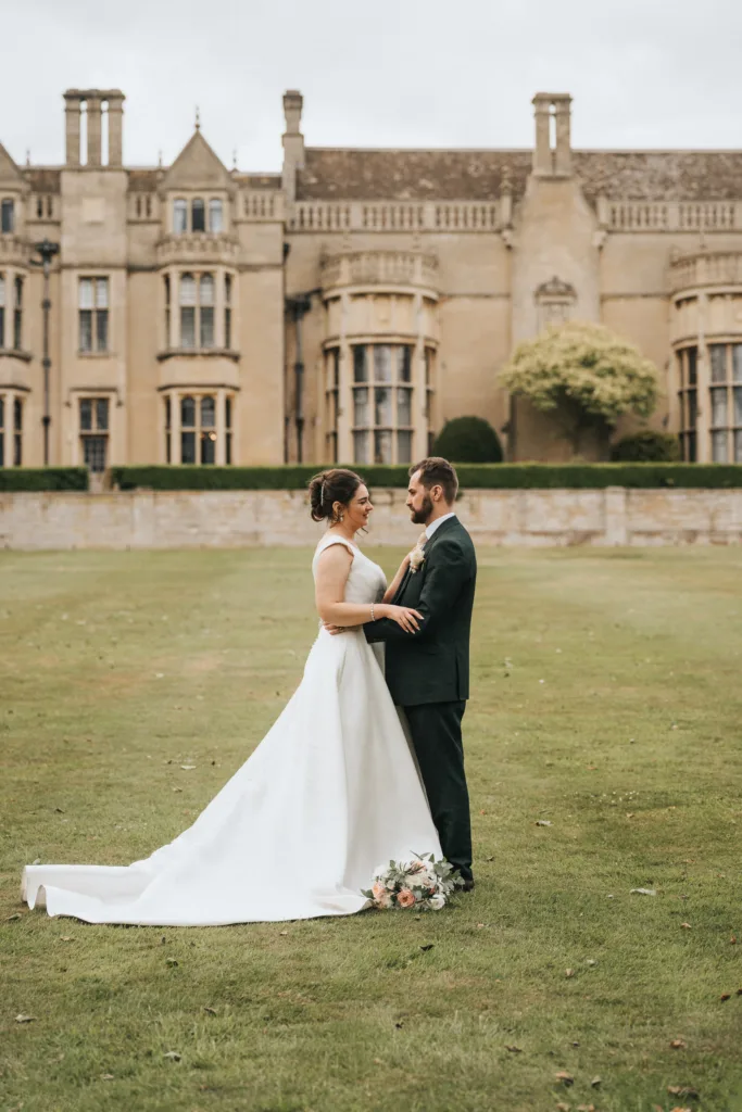 A bride in a long white dress and a groom in a dark suit stand facing each other, holding hands, on a grassy lawn in Kettering. A wedding photographer captures the moment by the historic stone building as a bouquet rests near the bride’s train. © Aimee Lince Photography - Wedding photographer in Lincolnshire, Yorkshire & Nottinghamshire