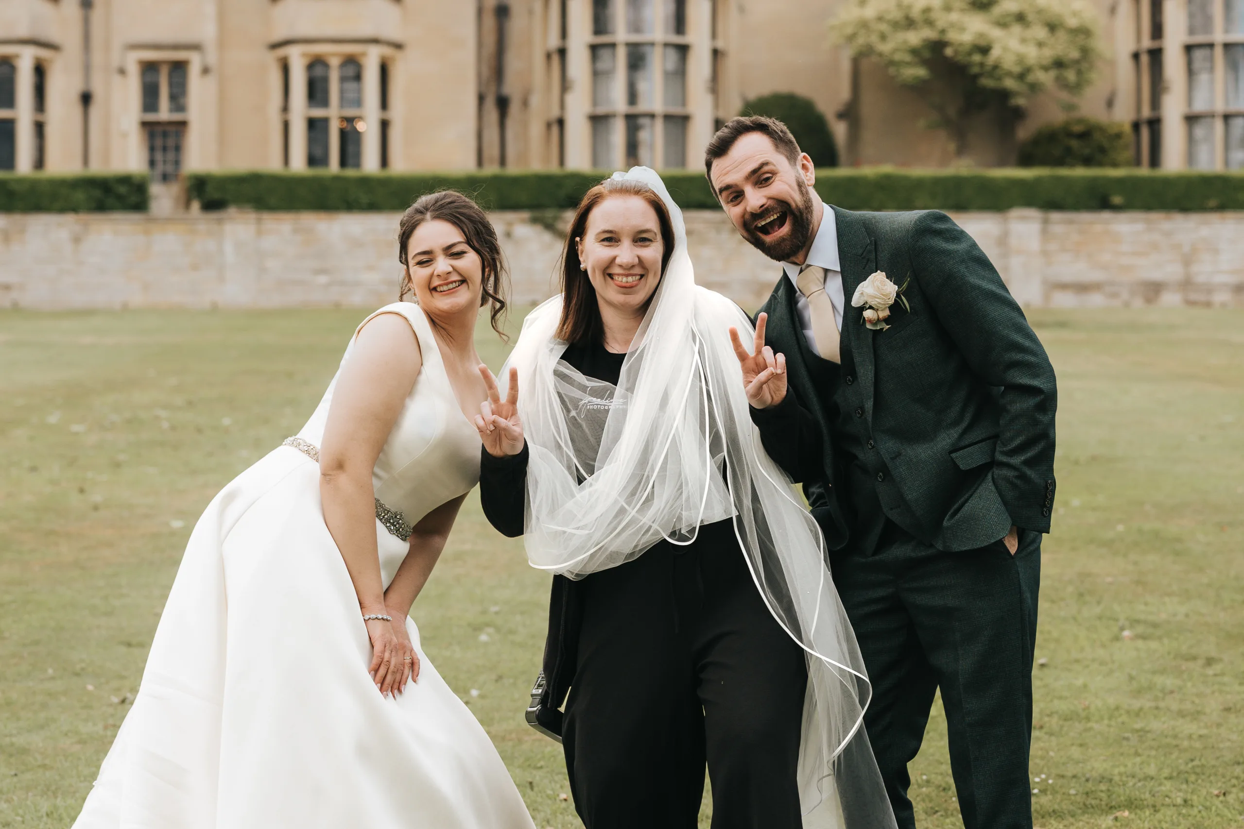 katie-adair-wedding-photography-rushton-hall-kettering-northamptonshire-721-of-1141-©-aimee-lince-photography-05-06-2025 Three adults pose happily on a lawn—a bride in white, a man in a dark green suit, and a woman in black flashing peace signs. A historic building sits behind them. This candid wedding photography was captured by an East Midlands wedding photographer. © Aimee Lince Photography - Wedding photographer in Lincolnshire, Yorkshire & Nottinghamshire