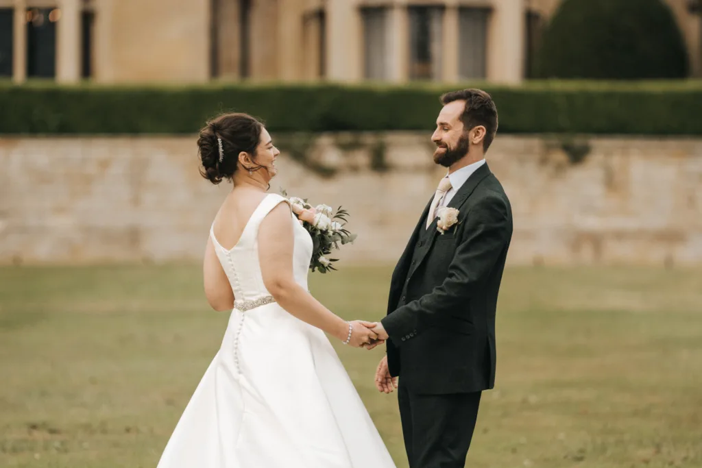 A bride in a white dress holding a bouquet smiles at her groom in a dark suit as they hold hands outdoors on the grassy lawn of Rushton Hall, with its stone wall and hedges softly blurred in the background of this Kettering wedding. © Aimee Lince Photography - Wedding photographer in Lincolnshire, Yorkshire & Nottinghamshire