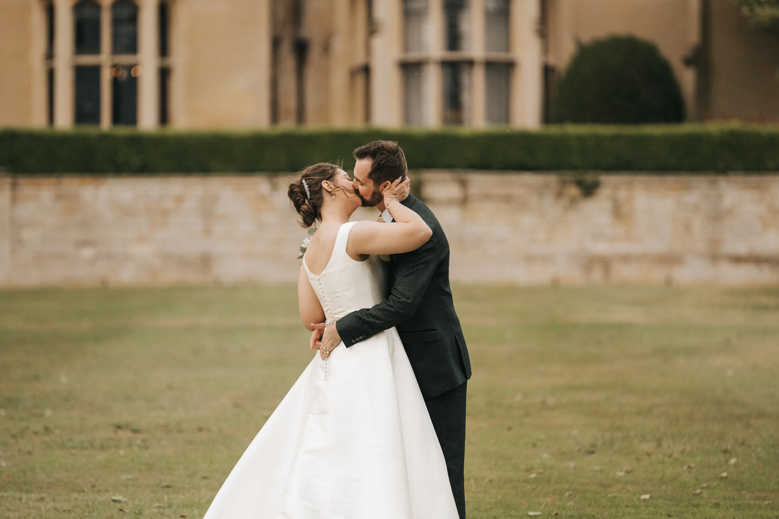 katie-adair-wedding-photography-rushton-hall-kettering-northamptonshire-731-of-1141-©-aimee-lince-photography-05-06-2025 A bride in a white gown and groom in a dark suit share a kiss outdoors on a lawn at Rushton Hall, with manicured hedges and elegant windows softly blurred behind them. Captured by a fun wedding photographer UK, they embrace closely as the bride’s back faces the camera. © Aimee Lince Photography - Wedding photographer in Lincolnshire, Yorkshire & Nottinghamshire