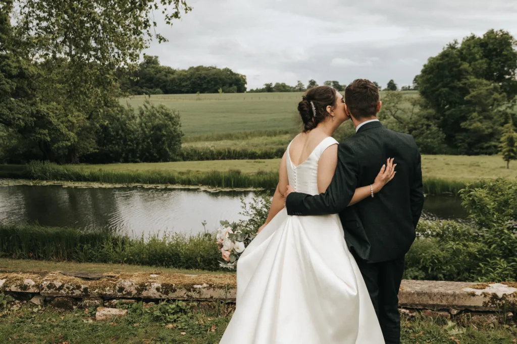 A bride in a white dress and groom in a dark suit stand arm in arm, gazing at a pond and green landscape. Captured by a Kettering wedding photographer, they face away from the camera amid trees and fields under a cloudy sky. © Aimee Lince Photography - Wedding photographer in Lincolnshire, Yorkshire & Nottinghamshire