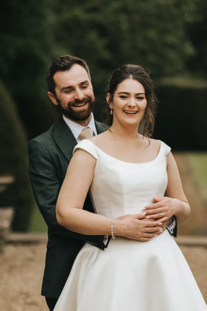 A smiling bride in an off-shoulder white wedding dress stands in front of her groom, who hugs her from behind. Captured by a wedding photographer in Kettering, both appear happy and relaxed outdoors with greenery and blurred trees behind them. © Aimee Lince Photography - Wedding photographer in Lincolnshire, Yorkshire & Nottinghamshire
