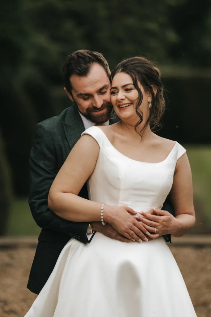 A groom in a dark suit lovingly embraces a smiling bride in a white satin gown outdoors at Rushton Hall near Kettering. The bride’s hair is styled up, with pearl earrings and bracelet. Both look happy amid softly blurred greenery in the background. © Aimee Lince Photography - Wedding photographer in Lincolnshire, Yorkshire & Nottinghamshire