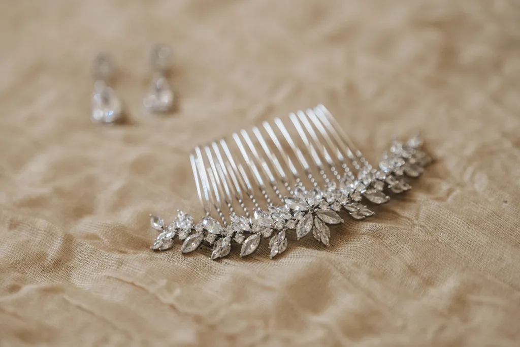 A silver hair comb adorned with sparkling, leaf-shaped crystals rests on textured beige fabric. In the blurred background, teardrop crystal earrings appear—perfect details for candid wedding photography or a stunning Rushton Hall wedding. © Aimee Lince Photography - Wedding photographer in Lincolnshire, Yorkshire & Nottinghamshire