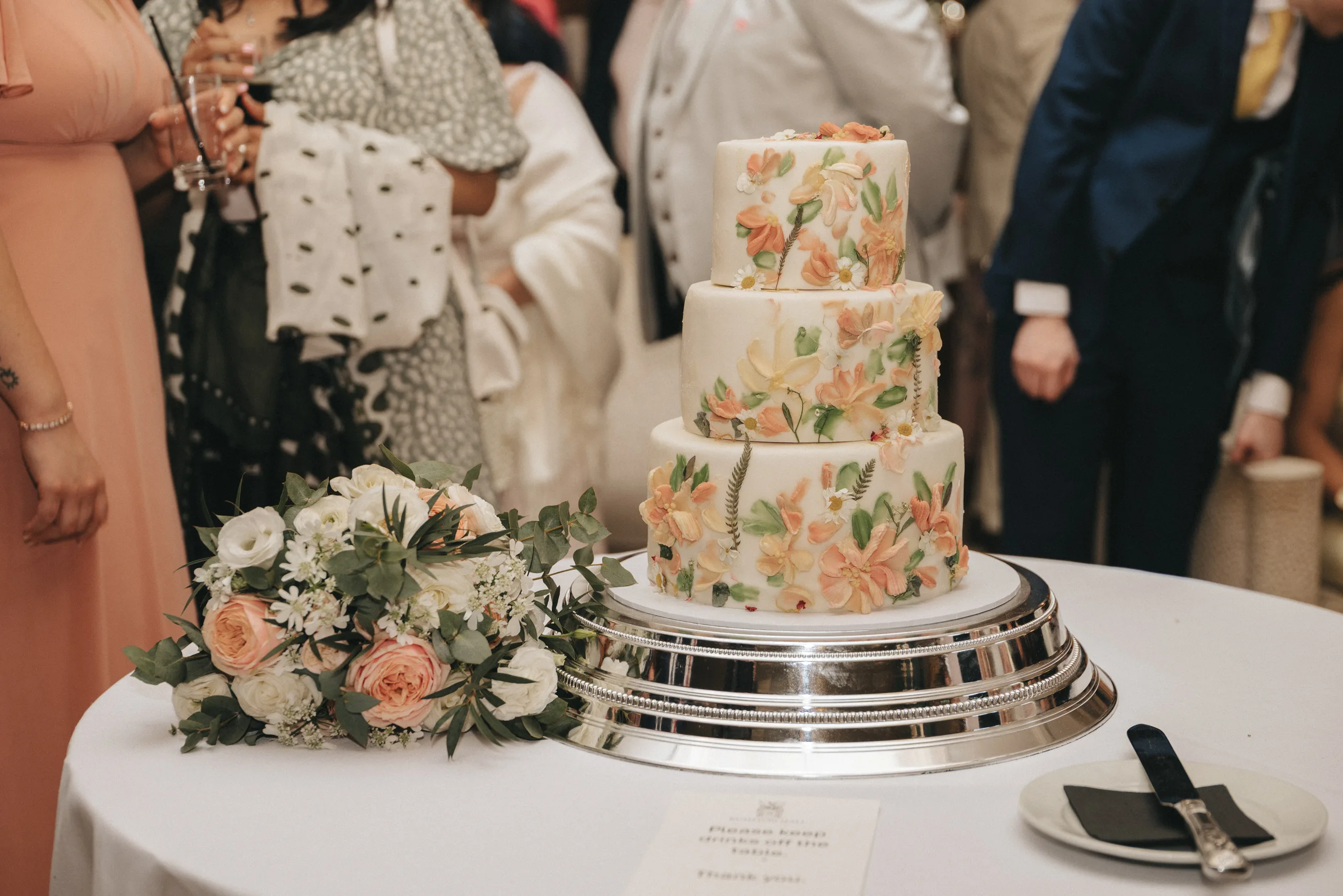 katie-adair-wedding-photography-rushton-hall-kettering-northamptonshire-763-of-1141-©-aimee-lince-photography-05-06-2025 A three-tiered white wedding cake with hand-painted pastel flowers and greenery sits on a silver stand at Rushton Hall. A bouquet of white and pink flowers lies nearby as the photographer captures guests in formal attire gathered around. © Aimee Lince Photography - Wedding photographer in Lincolnshire, Yorkshire & Nottinghamshire