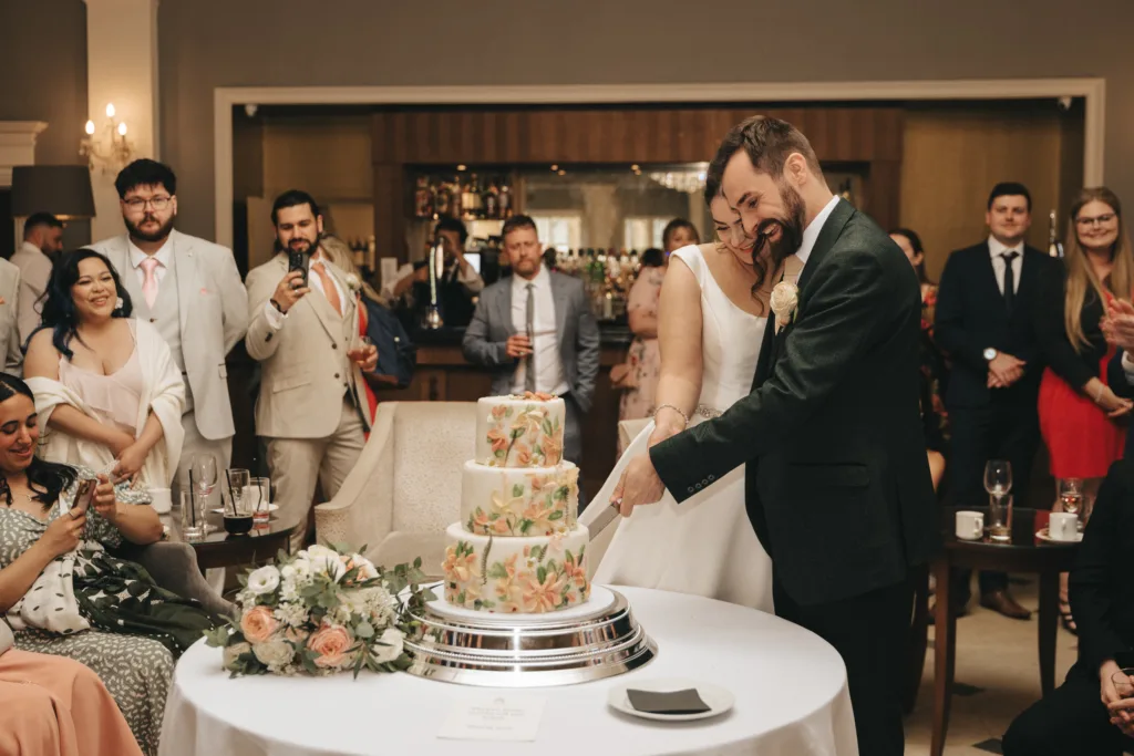 A bride and groom, smiling, cut a three-tiered floral wedding cake at Rushton Hall in a warmly lit room. Guests in formal attire watch as the photographer captures the joyful, celebratory moment; bouquets and drinks rest on nearby tables. © Aimee Lince Photography - Wedding photographer in Lincolnshire, Yorkshire & Nottinghamshire