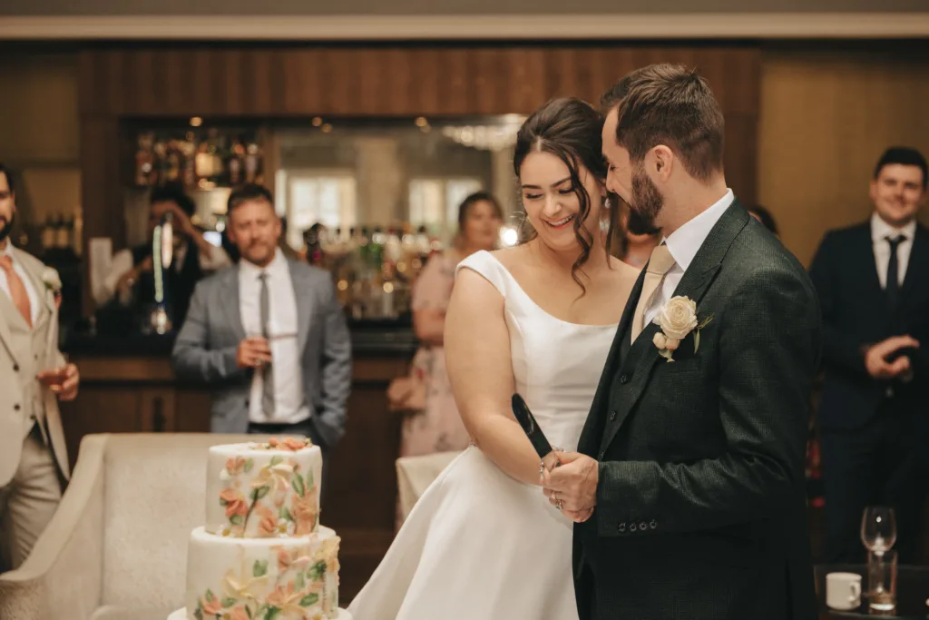 A bride and groom stand smiling together at Rushton Hall, holding a knife to cut their floral-decorated wedding cake. The photographer captures the joyful moment as guests in formal attire watch and smile in the background. © Aimee Lince Photography - Wedding photographer in Lincolnshire, Yorkshire & Nottinghamshire