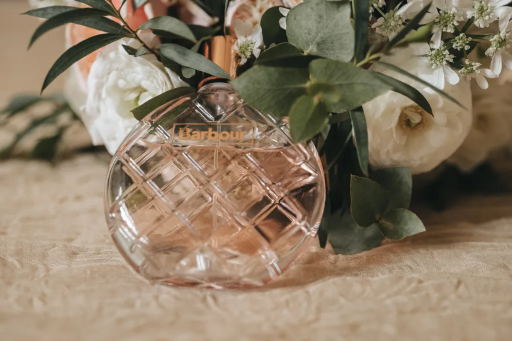 A round, pink, textured glass perfume bottle labeled "Barbour" sits on a beige fabric surface. Green leaves and white flowers surround it, creating a soft, elegant scene—perfect inspiration for a Kettering wedding photographer seeking natural beauty. © Aimee Lince Photography - Wedding photographer in Lincolnshire, Yorkshire & Nottinghamshire