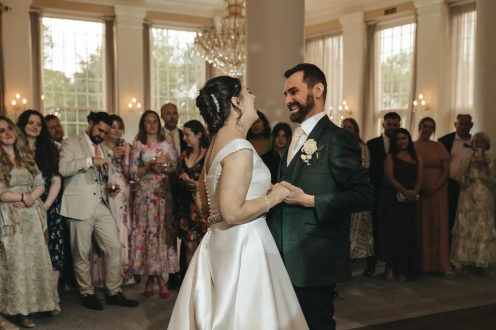 A bride and groom smile and dance together in a bright, elegant room at Rushton Hall, surrounded by wedding guests. The photographer captures the bride’s white gown and the groom’s boutonniere as guests watch near large windows. © Aimee Lince Photography - Wedding photographer in Lincolnshire, Yorkshire & Nottinghamshire