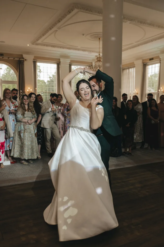 A joyful bride in a white gown and her smiling groom dance energetically at their wedding in the elegant Rushton Hall near Kettering, surrounded by guests in formal attire under sparkling chandeliers. © Aimee Lince Photography - Wedding photographer in Lincolnshire, Yorkshire & Nottinghamshire