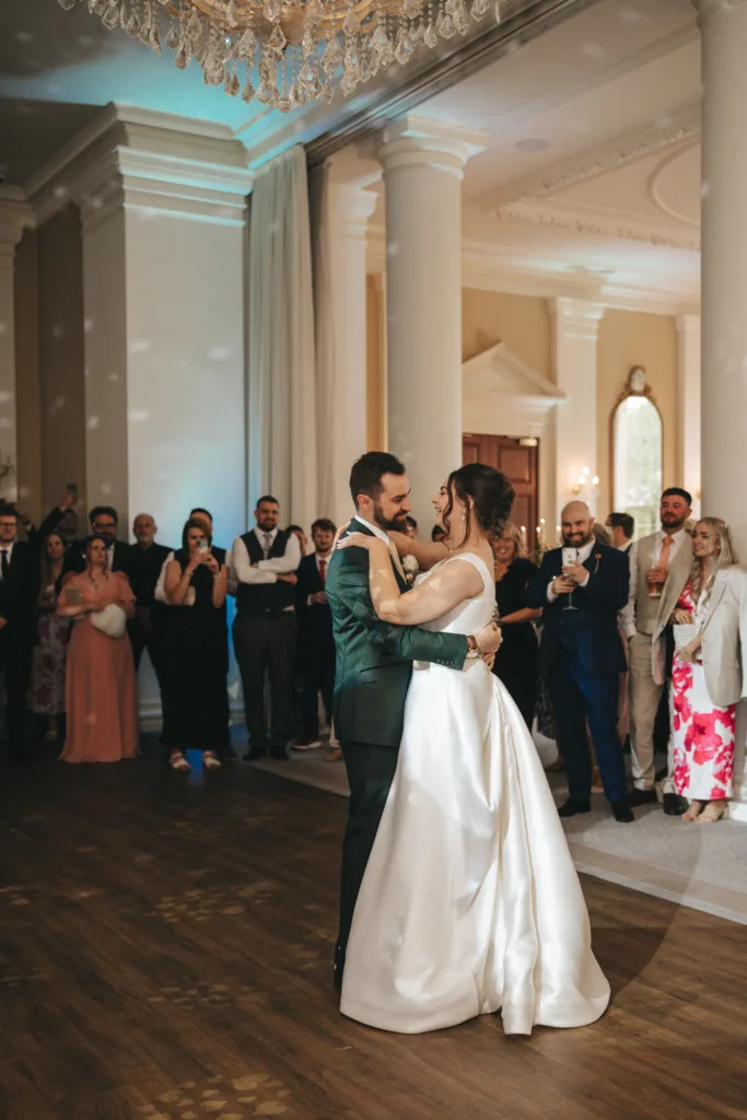 A bride in a white gown and a groom in a dark suit share their first dance at their Rushton Hall wedding. Guests in formal attire smile and capture the moment, while the photographer documents this elegant scene bathed in natural light through tall windows. © Aimee Lince Photography - Wedding photographer in Lincolnshire, Yorkshire & Nottinghamshire
