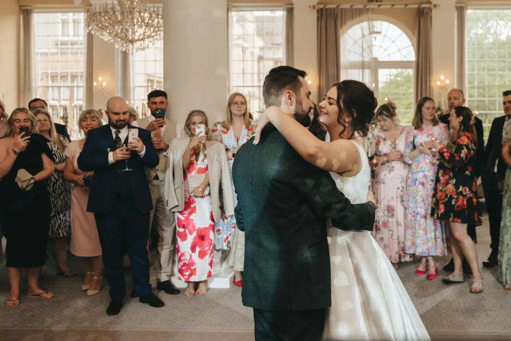 A bride and groom share their first dance in an elegant, light-filled room as a wedding photographer captures the moment. Large windows, chandeliers, and floral dresses create a stunning backdrop for this Kettering celebration. © Aimee Lince Photography - Wedding photographer in Lincolnshire, Yorkshire & Nottinghamshire