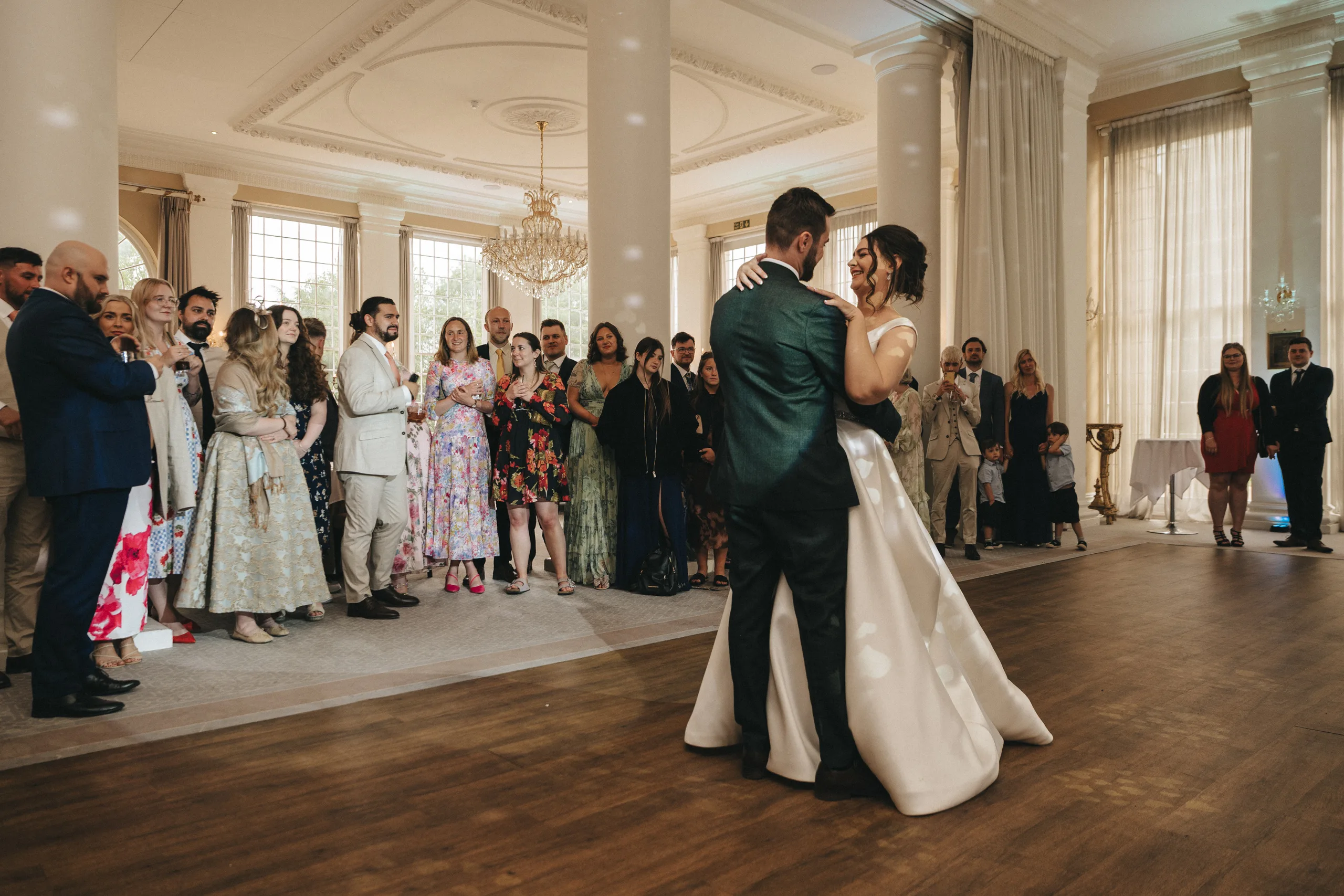 katie-adair-wedding-photography-rushton-hall-kettering-northamptonshire-795-of-1141-©-aimee-lince-photography-05-06-2025 A bride and groom share their first dance in the elegant Rushton Hall, with a wedding photographer capturing the moment. Guests stand in a semi-circle, smiling as warm light fills the grand hall’s tall columns and large windows. © Aimee Lince Photography - Wedding photographer in Lincolnshire, Yorkshire & Nottinghamshire