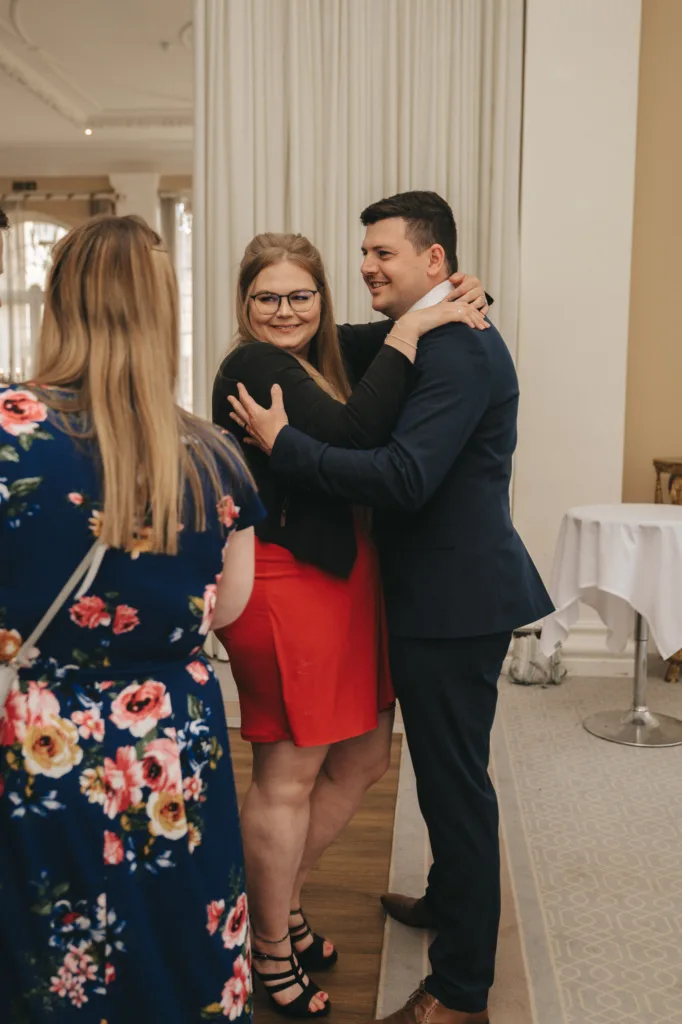 A man in a navy suit and a woman in a red dress and black top embrace and smile at the camera. Another woman in a blue floral dress stands nearby with her back facing the camera. The setting is indoors with neutral décor and a round table visible. © Aimee Lince Photography - Wedding photographer in Lincolnshire, Yorkshire & Nottinghamshire