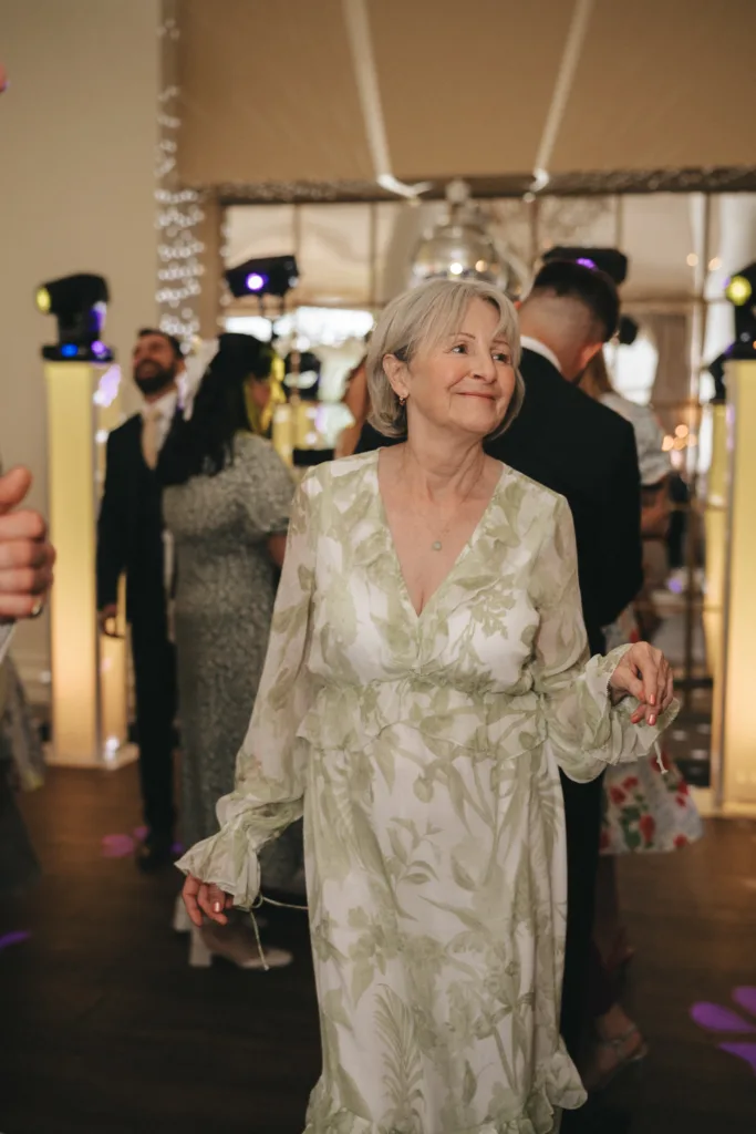 An older woman with short gray hair, wearing a long, flowing, light green and white floral dress, smiles as she stands in a warmly lit room. Other people are visible in the blurred background, suggesting a festive or social event. © Aimee Lince Photography - Wedding photographer in Lincolnshire, Yorkshire & Nottinghamshire