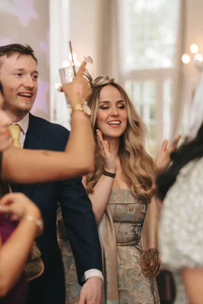 A smiling man in a blue suit and a woman with long blonde hair in a patterned dress stand in a bright room. The woman claps while people around them raise glasses, suggesting a celebratory event. The mood is joyful and festive. © Aimee Lince Photography - Wedding photographer in Lincolnshire, Yorkshire & Nottinghamshire