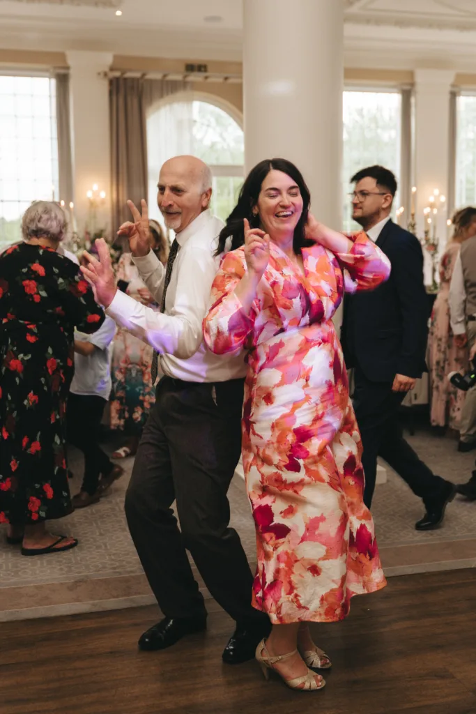 A smiling man in a white shirt and tie and a woman in a colorful floral dress dance back-to-back at a lively indoor event, with other guests in the background and elegant decor with large windows and candlelit tables visible. © Aimee Lince Photography - Wedding photographer in Lincolnshire, Yorkshire & Nottinghamshire