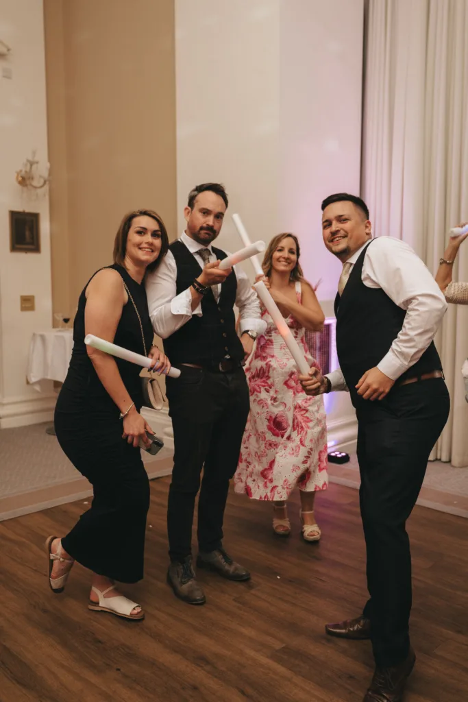 Four adults in semi-formal attire smile and pose playfully at an indoor event, holding white foam batons. Three stand in the foreground, two men in vests and a woman in a black dress, while another woman in a floral dress stands slightly behind them, also smiling. © Aimee Lince Photography - Wedding photographer in Lincolnshire, Yorkshire & Nottinghamshire