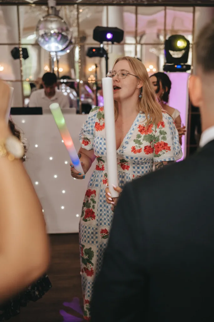 A woman with long blonde hair and glasses, wearing a floral dress, holds two glowing light sticks at a party. People surround her, and a DJ booth with lights and a disco ball is visible in the festive background. © Aimee Lince Photography - Wedding photographer in Lincolnshire, Yorkshire & Nottinghamshire