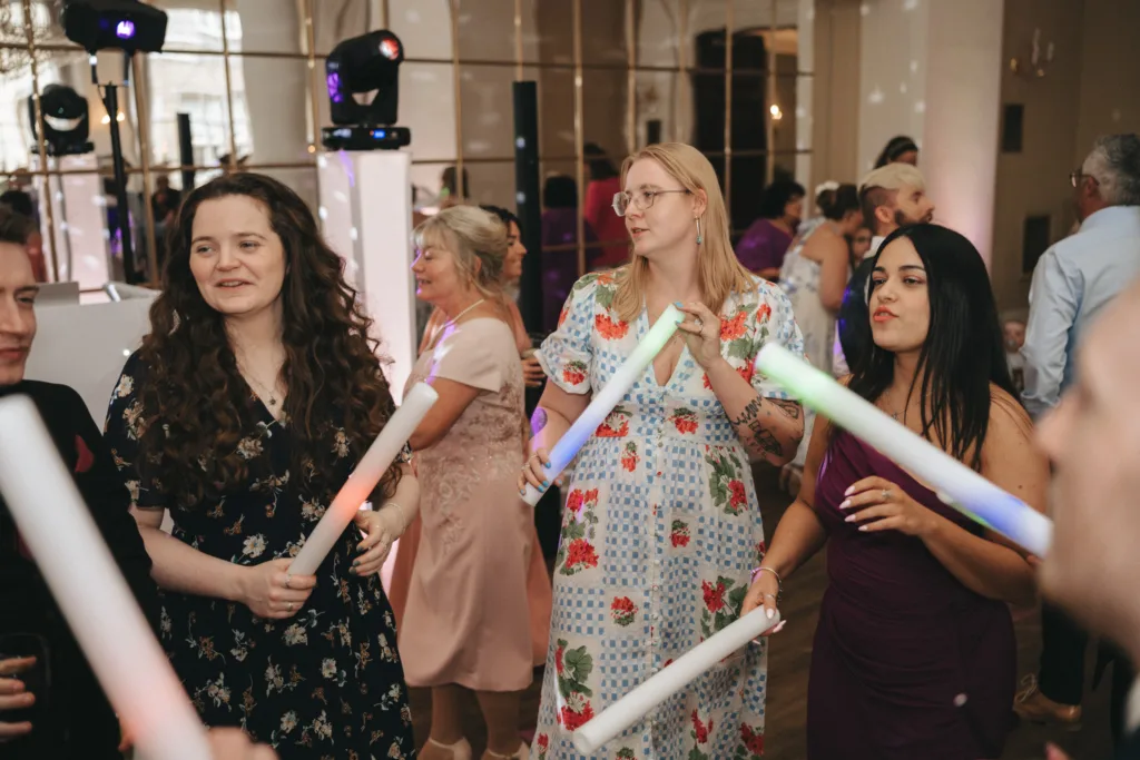Four women stand dancing and holding glowing tubes at an indoor party or wedding reception. They wear colorful dresses and appear to be enjoying themselves. Other guests and decorative lights are visible in the lively, festive background. © Aimee Lince Photography - Wedding photographer in Lincolnshire, Yorkshire & Nottinghamshire