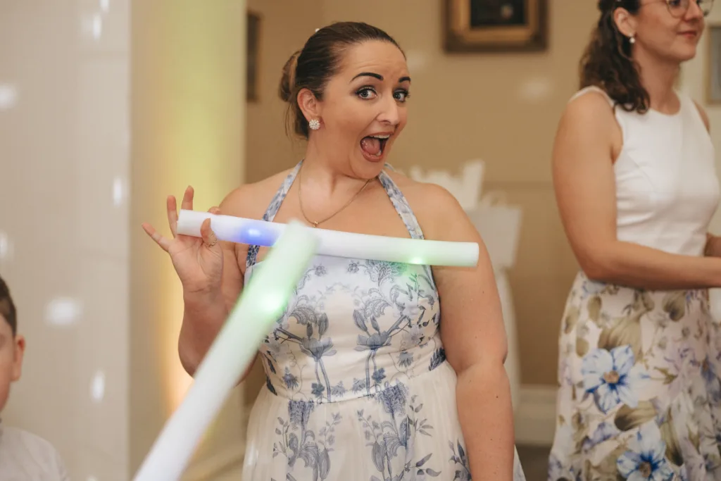 A woman in a white and blue floral dress smiles excitedly while holding a glowing light stick. She has her hair pulled back and is posing playfully. Another woman in a white dress with a floral skirt stands in the background, partially visible. © Aimee Lince Photography - Wedding photographer in Lincolnshire, Yorkshire & Nottinghamshire