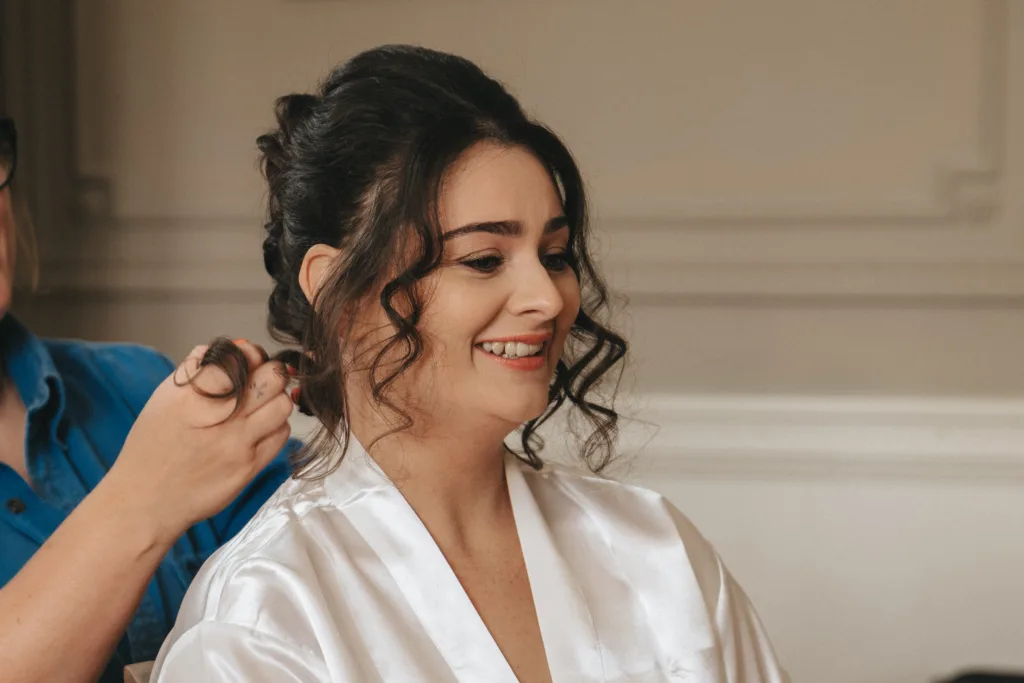 A woman with dark, curly hair sits smiling as another person styles her hair—she’s relaxed in a white satin robe. The neutral background hints at a calm indoor setting, perfect for a Rushton Hall wedding captured by a fun wedding photographer UK. © Aimee Lince Photography - Wedding photographer in Lincolnshire, Yorkshire & Nottinghamshire