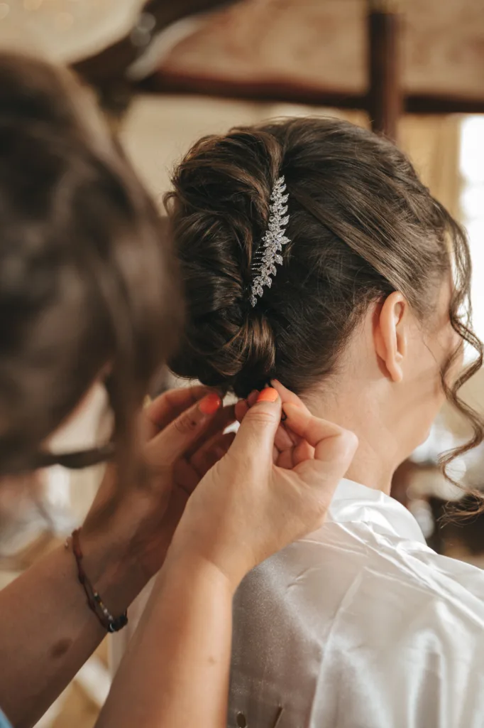 A person styles a woman's brown hair into an elegant updo, adorned with a silver, leafy accessory at her Rushton Hall wedding. Captured in candid wedding photography, she wears a white satin robe as the stylist adjusts her hair with both hands. © Aimee Lince Photography - Wedding photographer in Lincolnshire, Yorkshire & Nottinghamshire