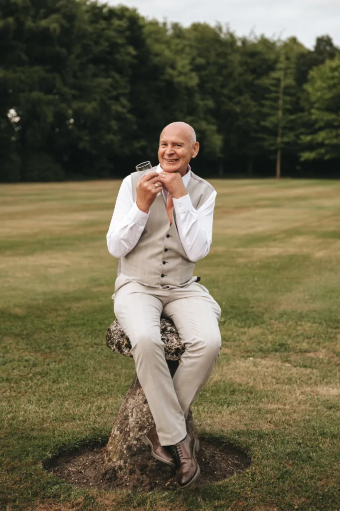 A bald man in a light grey vest and trousers, white shirt, and brown shoes sits on a stone in a grassy field, holding a glass and smiling, with trees in the background under a cloudy sky. © Aimee Lince Photography - Wedding photographer in Lincolnshire, Yorkshire & Nottinghamshire