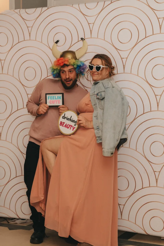 Two people pose playfully in front of a retro circular-patterned backdrop. The man wears a horned rainbow wig, holding a “Feelin Cute” sign. The woman in a peach dress and jean jacket lifts her leg, wearing heart-shaped sunglasses and holding a “drinking BEAUTY” sign. © Aimee Lince Photography - Wedding photographer in Lincolnshire, Yorkshire & Nottinghamshire
