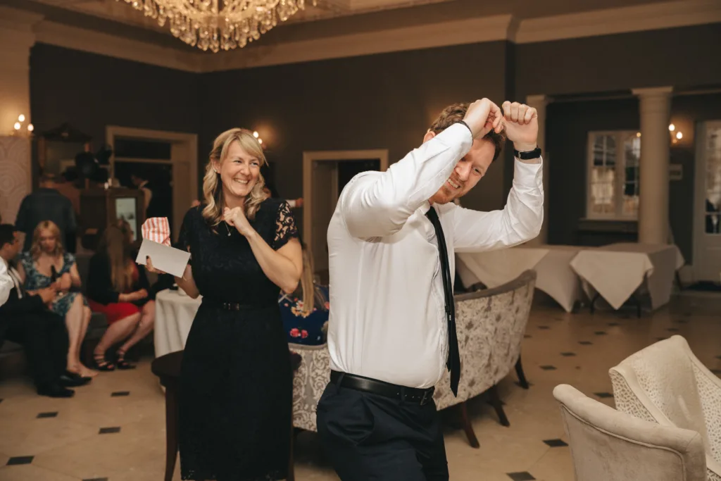 A smiling woman in a black lace dress holds an envelope while a man in a white shirt and tie dances playfully, arms up, in a warmly lit room with elegant décor; people are seated and chatting in the background. © Aimee Lince Photography - Wedding photographer in Lincolnshire, Yorkshire & Nottinghamshire