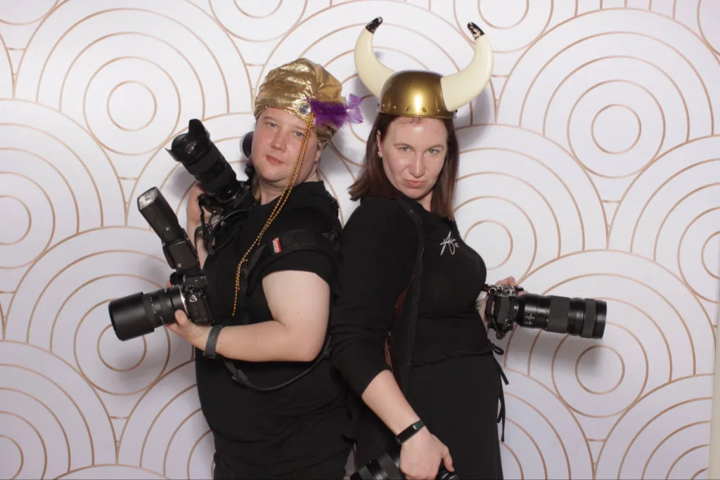 Two women in black clothes pose back-to-back, holding professional cameras. One wears a gold hat with a braid and feather, the other a Viking helmet with horns. Both have serious expressions. The background has a white and gold circular pattern. © Aimee Lince Photography - Wedding photographer in Lincolnshire, Yorkshire & Nottinghamshire