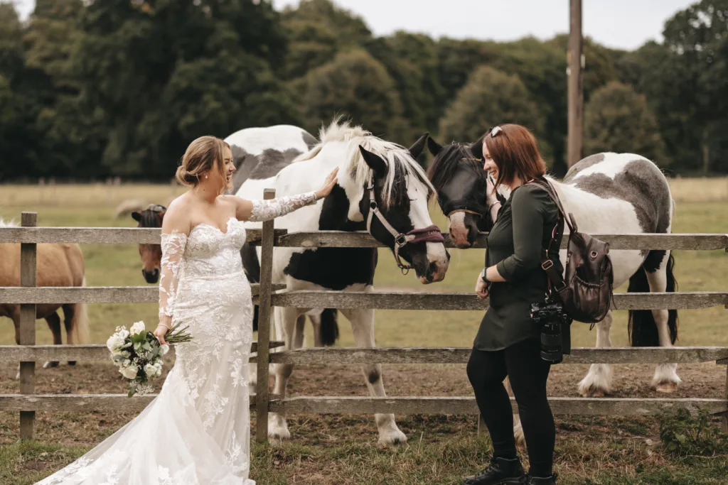 A bride in a white lace gown stands by a wooden fence, petting a black-and-white horse. She holds a bouquet and faces a woman in dark clothing with a camera bag. Two more horses and trees fill the rural background. © Aimee Lince Photography - Wedding photographer in Lincolnshire, Yorkshire & Nottinghamshire