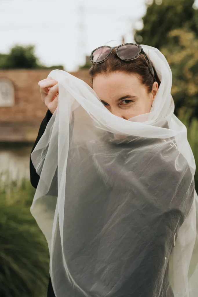 A person with brown hair pulled back and sunglasses on their head holds a sheer white fabric, partly covering their face. They gaze at the camera with only their eyes and forehead visible. The background is outdoors with greenery and a blurry building. © Aimee Lince Photography - Wedding photographer in Lincolnshire, Yorkshire & Nottinghamshire