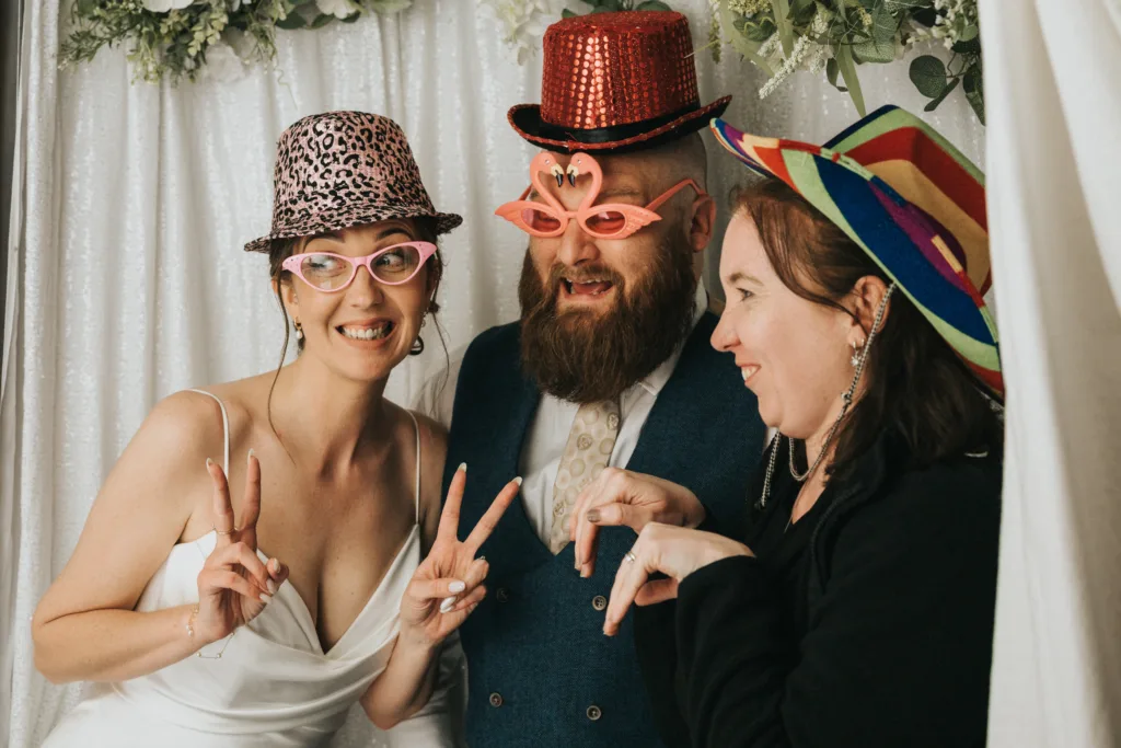 Three adults pose playfully in front of a white curtain. The woman on the left wears a white dress, pink leopard hat, and pink glasses. The man in the center wears a red sequined hat and flamingo glasses. The woman on the right wears a rainbow hat. All smile and gesture. © Aimee Lince Photography - Wedding photographer in Lincolnshire, Yorkshire & Nottinghamshire