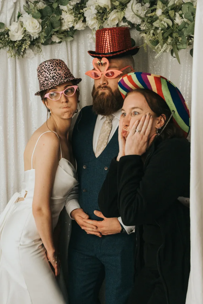 Three adults pose playfully in a photo booth with white floral decorations. One wears a white dress, pink leopard hat, and pink glasses; another, a blue suit, red hat, and flamingo glasses; the third wears a rainbow sombrero and holds her face, making an exaggerated expression. © Aimee Lince Photography - Wedding photographer in Lincolnshire, Yorkshire & Nottinghamshire