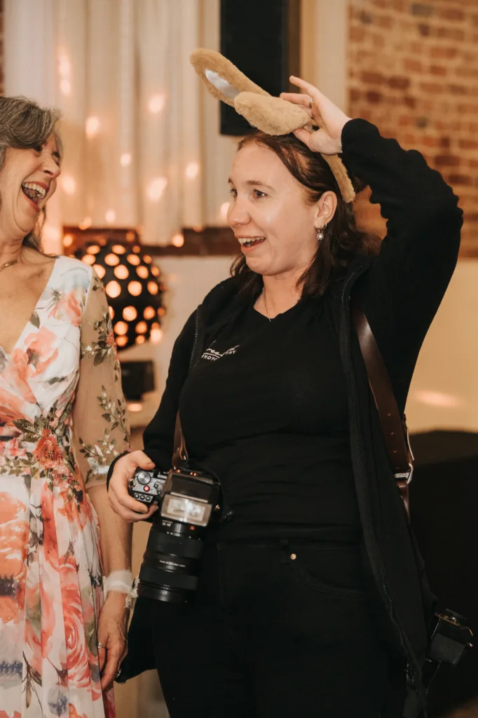 Two women smile and laugh indoors. The woman on the left wears a floral dress with sheer sleeves, while the woman on the right, holding a camera, wears black and playfully puts on tan bunny ears. Warm lighting and exposed brick add to the festive atmosphere. © Aimee Lince Photography - Wedding photographer in Lincolnshire, Yorkshire & Nottinghamshire