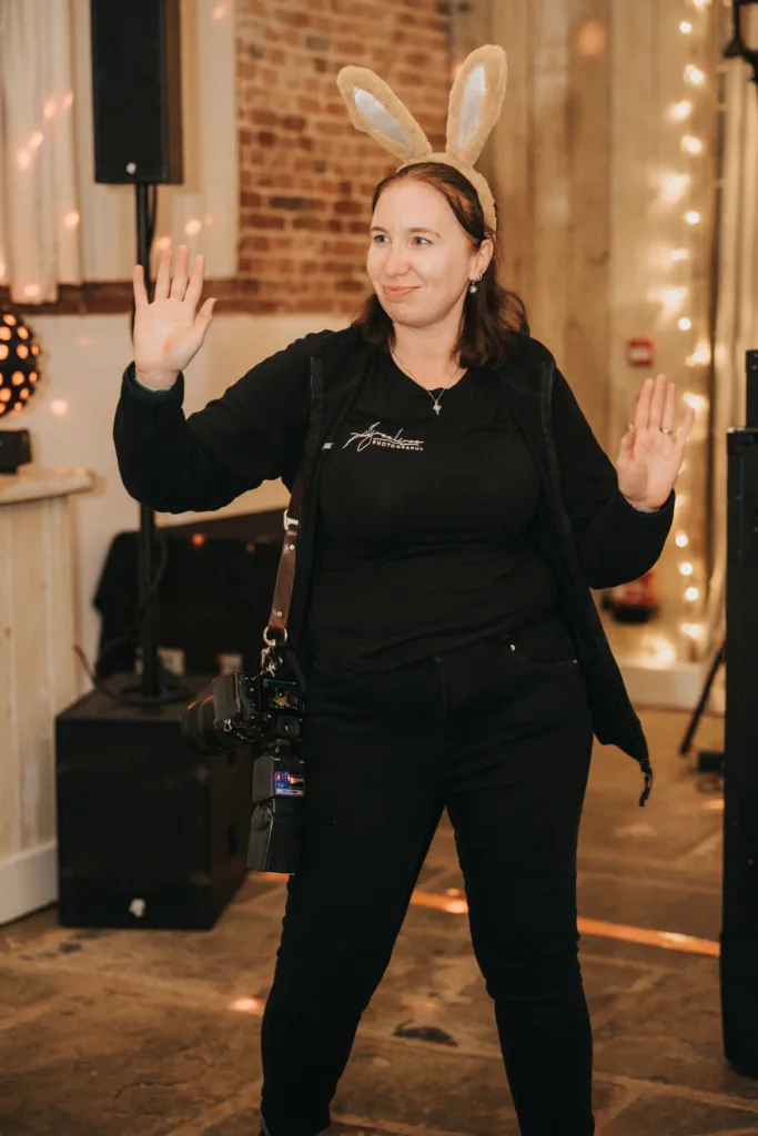 A woman wearing bunny ears and all-black clothing stands indoors with both hands raised. She has a camera hanging from her shoulder and is smiling. The background features exposed brick walls, string lights, and event equipment. © Aimee Lince Photography - Wedding photographer in Lincolnshire, Yorkshire & Nottinghamshire