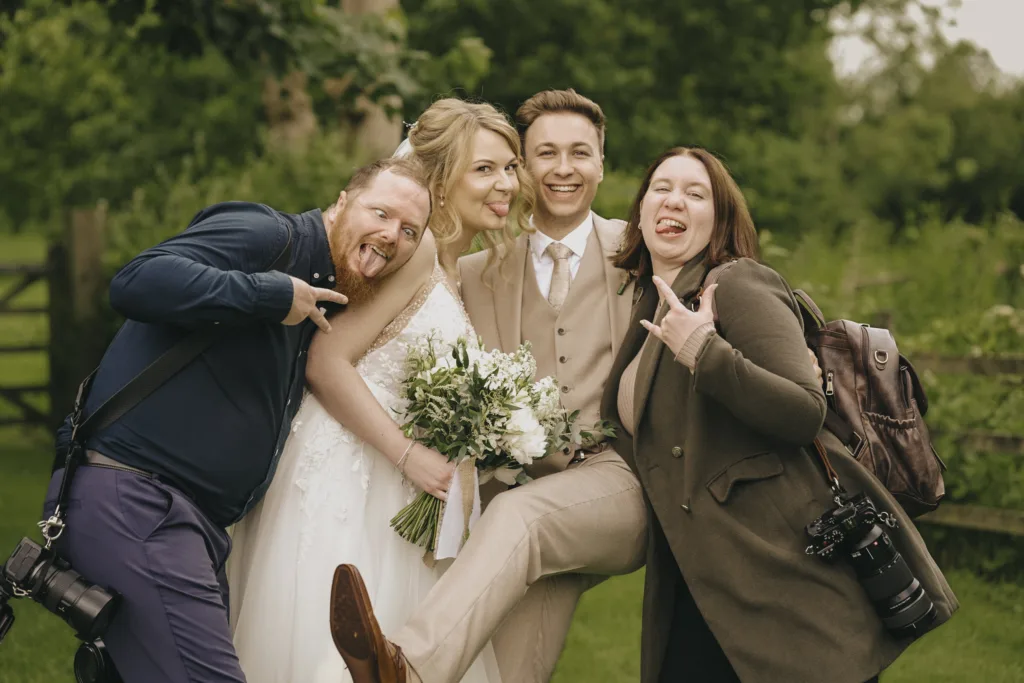Four people pose joyfully outdoors: a bride in a white gown holding a bouquet, a groom in a beige suit, and two casually dressed guests with cameras, making playful faces and gestures. Lush greenery and a wooden fence are in the background. © Aimee Lince Photography - Wedding photographer in Lincolnshire, Yorkshire & Nottinghamshire