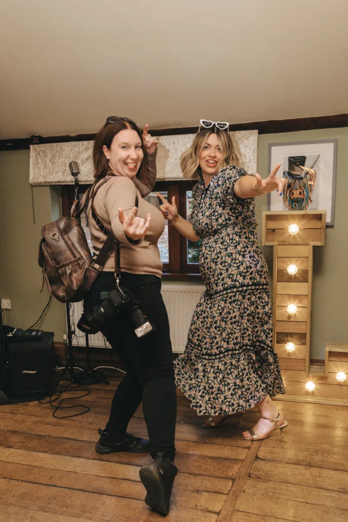 Two women enthusiastically pose indoors on a wooden floor. One wears a floral dress and sunglasses, while the other has a camera and backpack. Both smile and gesture energetically. Behind them are lights, a microphone, and art hanging on a green wall. © Aimee Lince Photography - Wedding photographer in Lincolnshire, Yorkshire & Nottinghamshire
