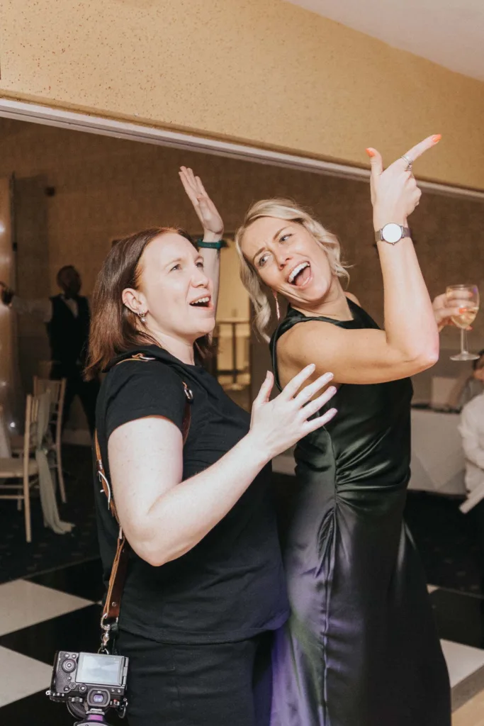 Two beautiful women smile and pose at a celebration. One, a photographer in black with a camera on her hip, stands next to a friend in a dark satin dress holding a wine glass. They enjoy the lively lifestyle on a black-and-white checkered floor. © Aimee Lince Photography - Wedding photographer in Lincolnshire, Yorkshire & Nottinghamshire