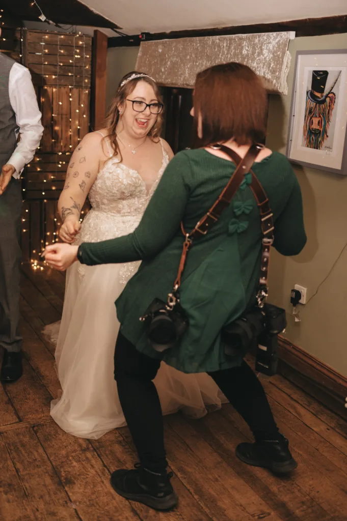 A smiling bride in a white dress and glasses holds hands and dances enthusiastically with a woman in a green cardigan who has two cameras strapped to her back. They are indoors on a wooden floor, surrounded by warm decorative lights. © Aimee Lince Photography - Wedding photographer in Lincolnshire, Yorkshire & Nottinghamshire