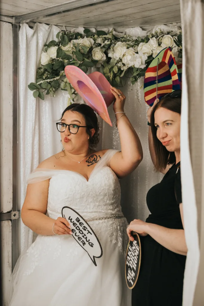 Two women pose in a photo booth with white floral decorations. One, in a wedding dress, lifts a pink hat and makes a playful face. The other, in a black dress, smiles. Both hold speech bubble props; one reads “Best wedding ever!”. © Aimee Lince Photography - Wedding photographer in Lincolnshire, Yorkshire & Nottinghamshire