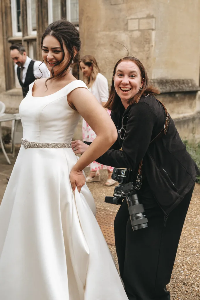 A smiling bride in a white dress stands outside an old stone building. Next to her, a laughing woman with cameras, possibly a photographer, helps adjust the dress. Two other people are blurred in the background, appearing cheerful. © Aimee Lince Photography - Wedding photographer in Lincolnshire, Yorkshire & Nottinghamshire