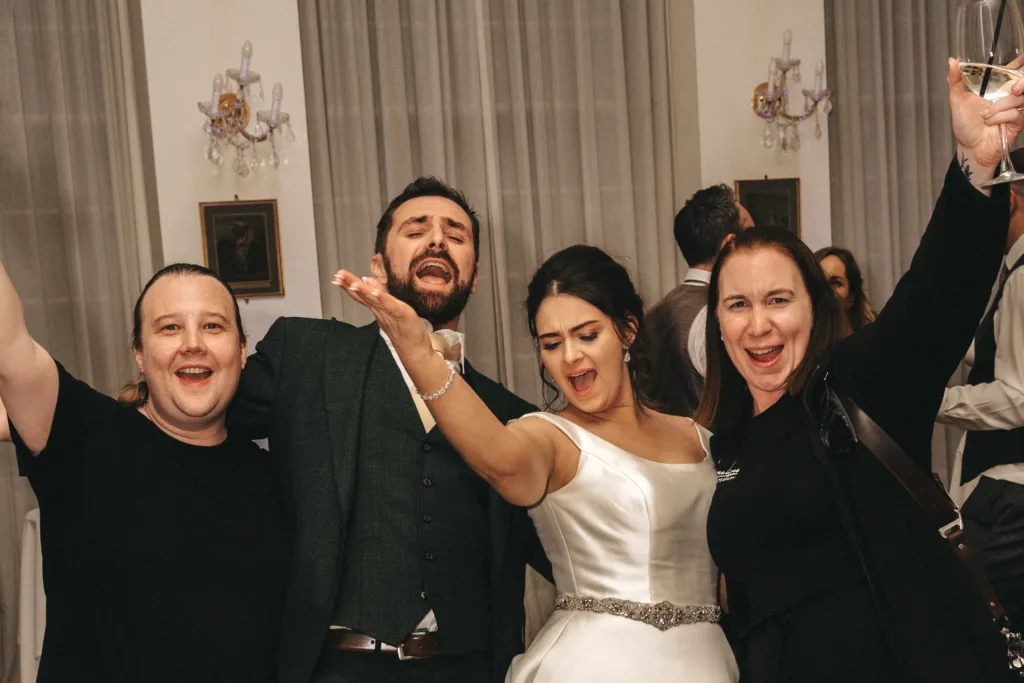 Four adults stand close together, joyfully posing and singing at a celebration. A bride in a white dress is between two men and another woman. Everyone has raised arms and happy expressions. Elegant decor, framed art, curtains, and soft lighting are in the background. © Aimee Lince Photography - Wedding photographer in Lincolnshire, Yorkshire & Nottinghamshire