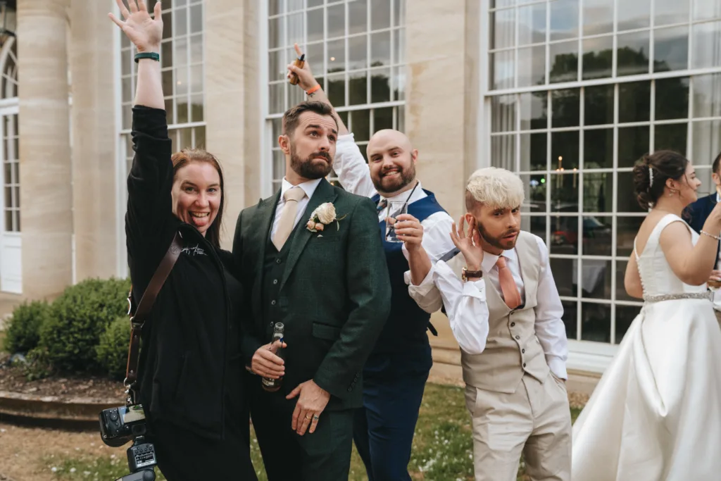 A group of four adults pose playfully outside a formal building. One woman raises her arm and smiles; a man in a green suit stands with a serious face; another man in a blue suit grins; a man in a beige vest hams it up. A bride is partially seen on the right. © Aimee Lince Photography - Wedding photographer in Lincolnshire, Yorkshire & Nottinghamshire
