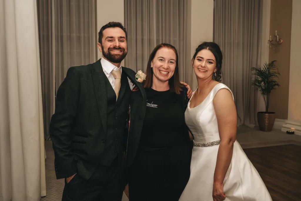 A smiling man in a dark suit and a beautiful woman in a white wedding dress stand on either side of a woman in black clothes. They are indoors with tall curtains and a plant, posing closely together for this signature lifestyle photo. © Aimee Lince Photography - Wedding photographer in Lincolnshire, Yorkshire & Nottinghamshire