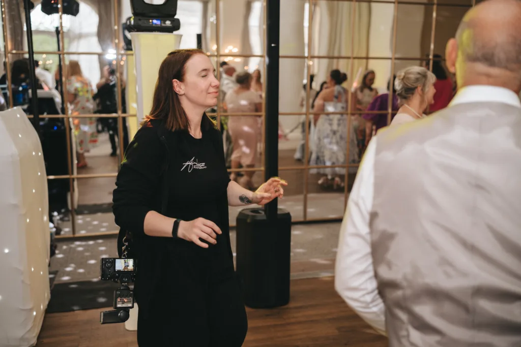 A woman in black with a camera and equipment belt smiles while dancing at an indoor event. People in formal attire and elegant dresses mingle in the background, and large mirrored panels and draped curtains decorate the room. © Aimee Lince Photography - Wedding photographer in Lincolnshire, Yorkshire & Nottinghamshire